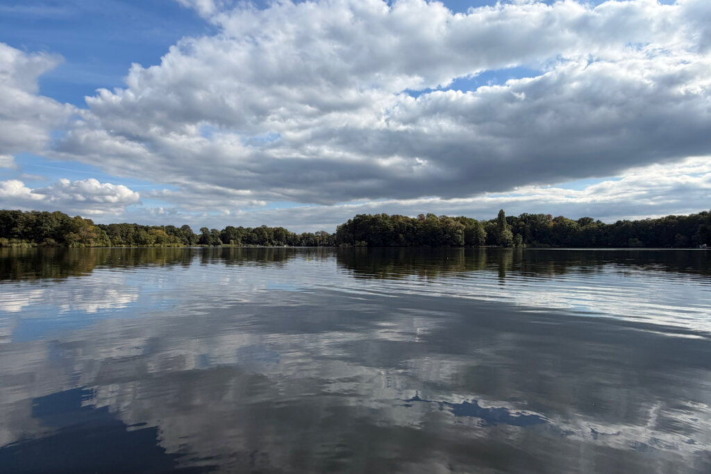Wolken spiegeln sich im glatten Wasser des Glindower Sees. Der See wird von einem Wald gesäumt.